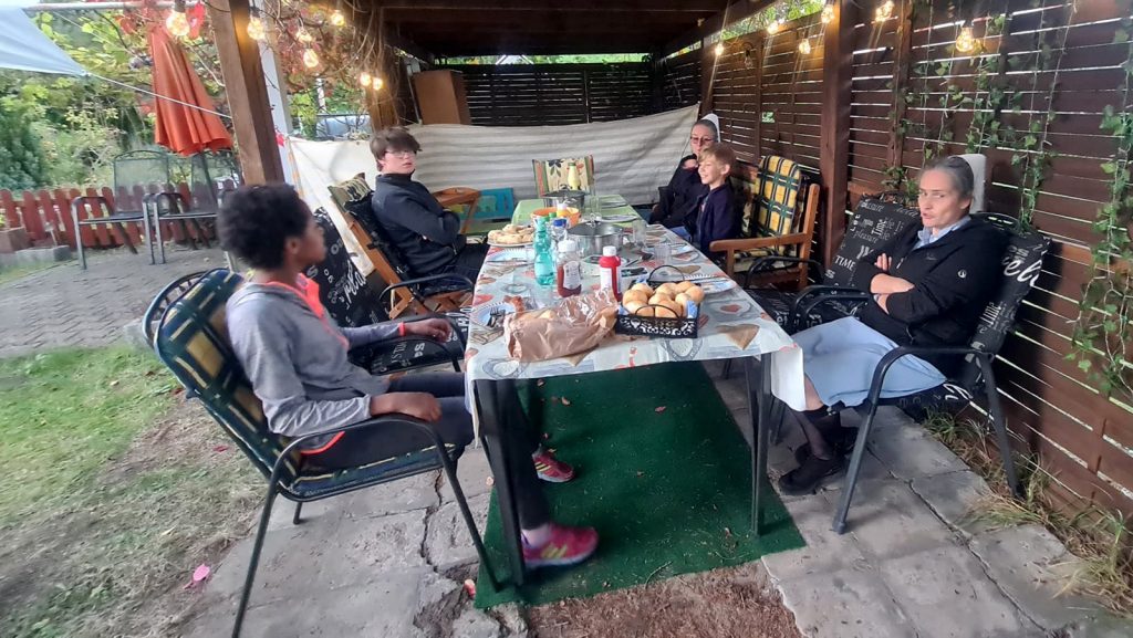 Grillveranstaltung im Garten des Kinderheim Elisabeth Stift. Kinder und Schwestern des Heims essen gemeinsam mit Mitgliedern des Vereins am Tisch in einer Gartenlaube.