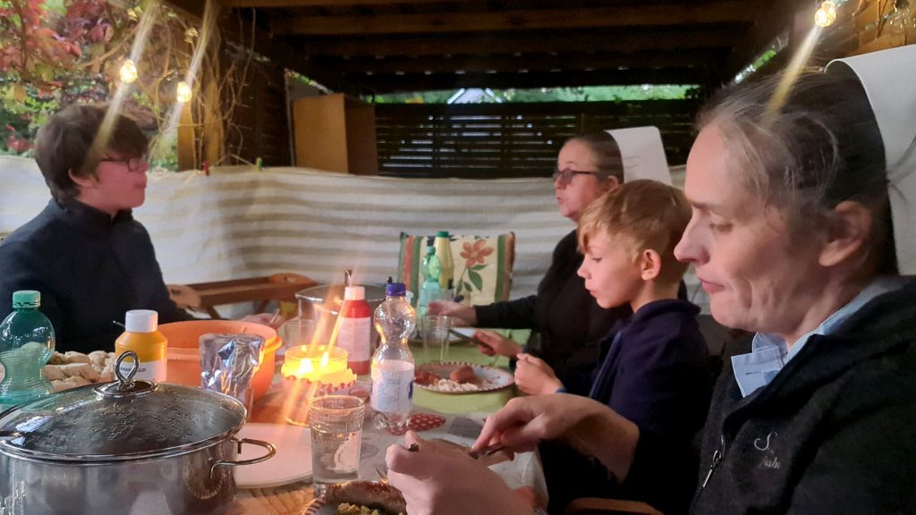 Grillveranstaltung im Garten des Kinderheim Elisabeth Stift. Kinder und Schwestern des Heims essen gemeinsam mit Mitgliedern des Vereins am Tisch in einer Gartenlaube.