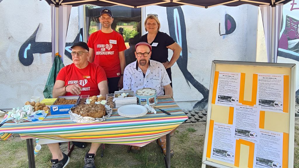 Vereinsmitglieder sitzen an ihrem Stand unter einem kleinen Pavillons. Der Tisch ist gedeckt mit Gebäck für Zuschauer und Sportler.
