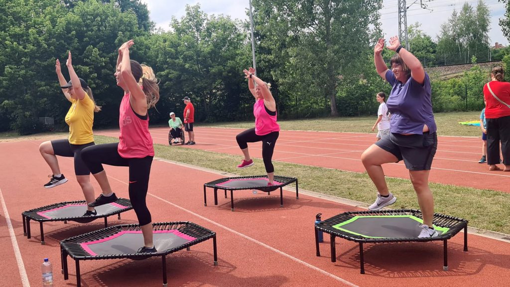Mehrere Damen üben gemeinsam auf Trampolins, die auf dem Sportplatz aufgestellt wurden. Vereinsmitglieder unterhalten sich mit verschiedenen Personen im Hintergrund. Auch ein Rollstuhlfahrer gehört zu den Zuschauern.