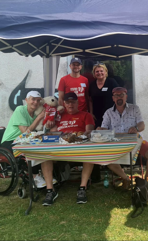 Vereinsmitglieder sitzen an ihrem Stand unter einem kleinen Pavillons. Der Tisch ist gedeckt mit Gebäck für Zuschauer und Sportler.