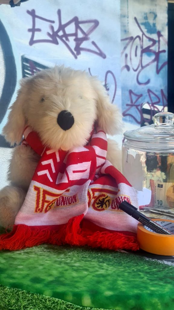 Ein weißfarbener Plüschhund mit Fan-Schal des 1. FC Union. Neben ihm steht ein Spendenglas.